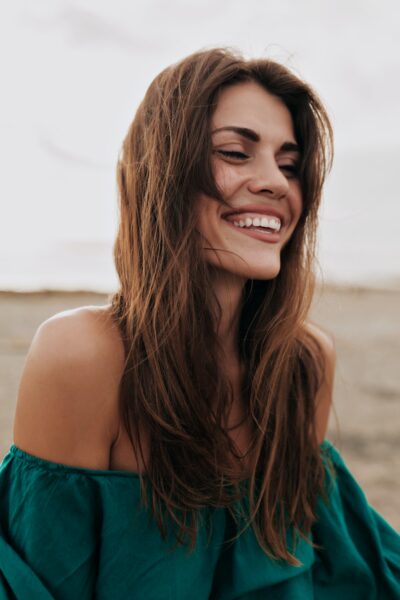 Happy charming model wearing dress with bare shoulders smiling with closed eyes on the beach